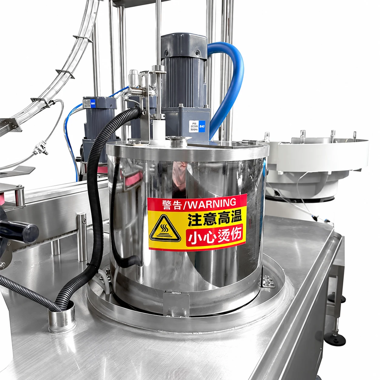 Stainless product supply tank with geared motor, hoses and a hot-surface warning label on a dosing-cap line